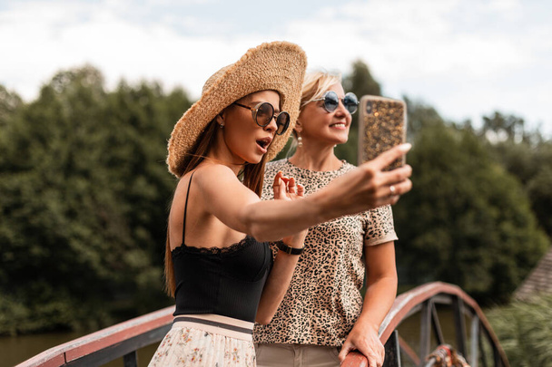 две красивые смешные женщины счастливые мама и дочь в модных солнцезащитных очках в шляпе и стильном летнем наряде сделать совместное фото селфи на смартфоне на отдыхе - Фото, изображение