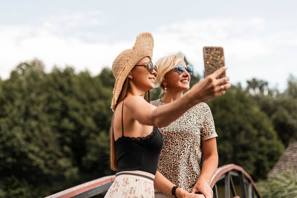 beautiful young woman with a smile and an elderly happy mom in sunglasses in fashionable summer clothes do a joint selfie photo and relax on sunny weekend - Foto, immagini