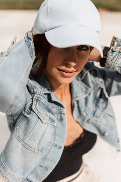 Moda serio bella giovane donna modello in elegante cappello con giacca di jeans con lingerie di pizzo si siede sulla sabbia bianca alla luce del sole - Foto, immagini