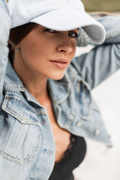 Cool stylish portrait of a beautiful young woman with a sexy look in a fashion blue cap and a blue denim jacket with lace tank top sits on the white sand - Photo, Image