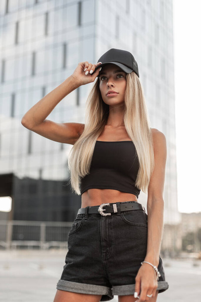 Fashion beautiful woman in a black stylish T-shirt and black jeans shorts with a cap walks in the city - Fotó, kép