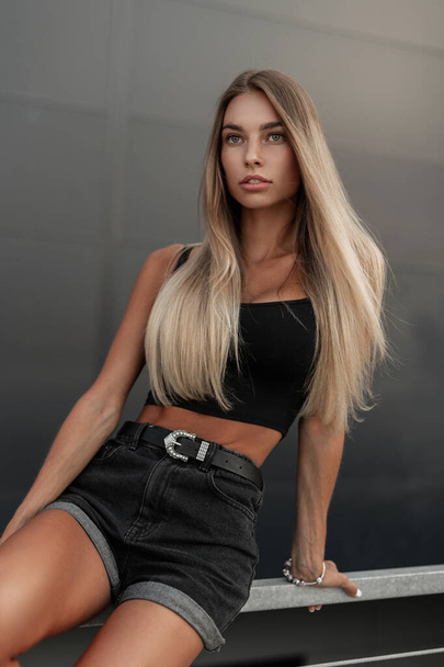 Pretty fashion model woman with beautiful body in black jeans shorts and black top t-shirt sits in the city - Fotó, kép