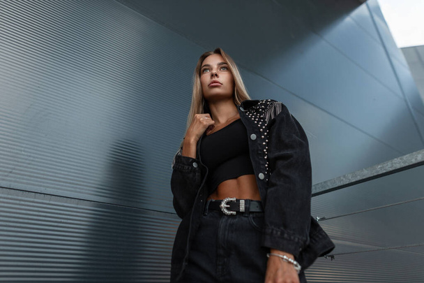 Swag mulher moda hipster com moda jaqueta de ganga preta em jeans elegantes ficar perto de parede de metal moderno na rua - Foto, Imagem
