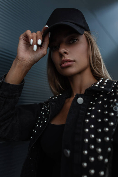 Όμορφη γυναίκα μοντέλο μόδας με δροσερό μαύρο καπέλο σε μοντέρνα ρούχα μαύρο τζιν με μοντέρνο σακάκι θέτει σε εξωτερικούς χώρους - Φωτογραφία, εικόνα