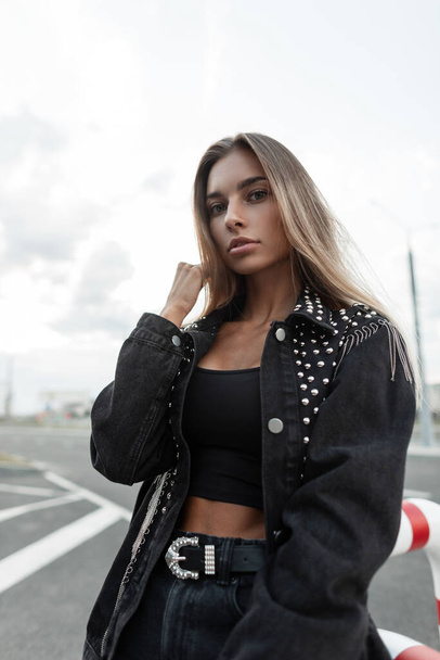 Beautiful young woman model with blonde hair with a pretty face in a fashion black denim jacket and a black top T-shirt with black jeans with a belt walks in the city - Photo, Image