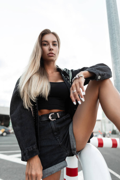 Glamorous beautiful young girl with blonde hair and shapely sexy legs with jeans shorts in a fashionable denim jacket and tank top poses in the city - Foto, afbeelding