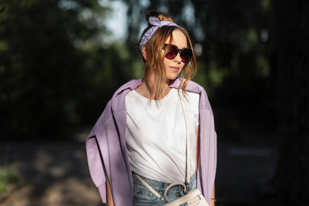 Bella giovane donna hipster con occhiali da sole di moda e una bandana con una felpa viola cammina per strada - Foto, immagini