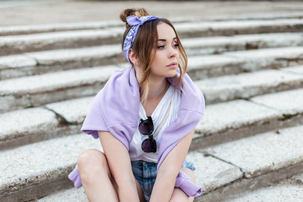Divatos hipszter nő, kendővel, fehér pólóban, napszemüveggel és lila kapucnival, kék farmernadrágban ül az utcán. - Fotó, kép