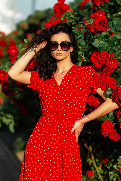 Kırmızı elbiseli, moda güneş gözlüklü zarif kadın parkta kırmızı güllerin yanında duruyor. - Fotoğraf, Görsel
