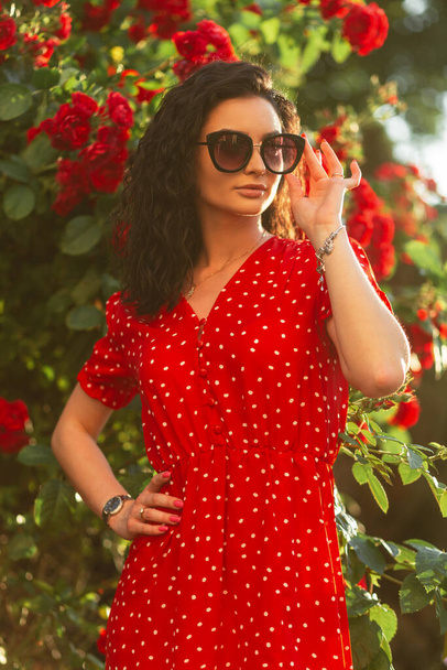 Jeune jolie femme élégante avec des lunettes de soleil de mode en robe rouge à pois pose près des roses au coucher du soleil - Photo, image
