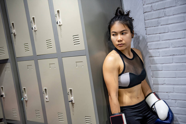Joven mujer asiática boxeadora con guantes de boxeo de pie junto al armario en el vestuario. Clase de boxeo femenino o concepto de artes marciales - Foto, Imagen