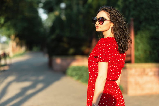 Divatos nő elegáns, elegáns, vörös ruhában, napszemüveggel, aki az utcán sétál naplementekor. - Fotó, kép