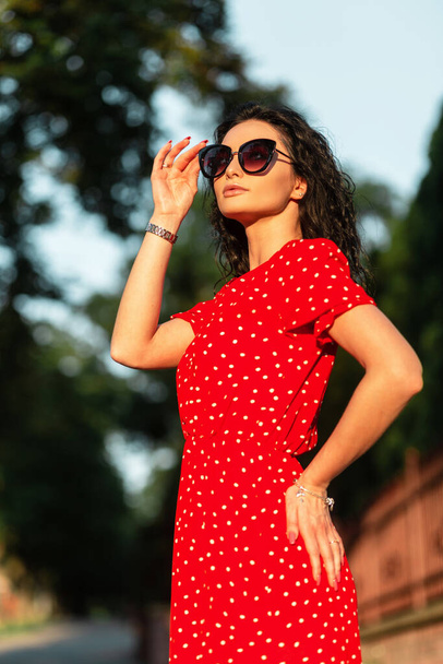 Κομψότητα όμορφη γυναίκα βάζει γυαλιά ηλίου μόδας σε vintage κόκκινο φόρεμα με τα πόδια στο δρόμο στο ηλιοβασίλεμα - Φωτογραφία, εικόνα