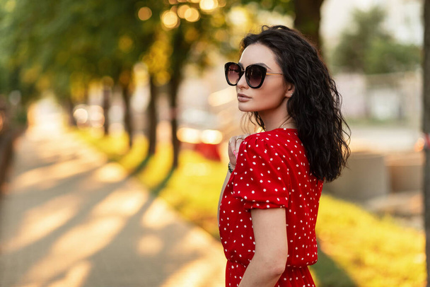 Fashion young hipster woman with sunglasses in red fashionable dress walking on the street at sunset - Foto, imagen