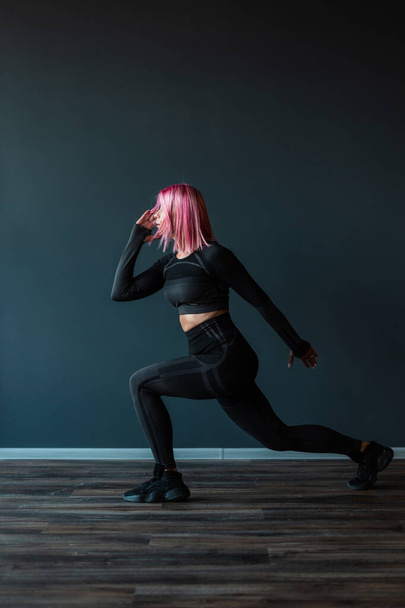 Hermosa mujer joven fitness con el pelo rosa en ropa deportiva negro preparándose para correr cerca de la pared negra - Foto, Imagen
