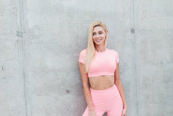 Pretty smiling fitness model woman with blond hair in fashion pink sportswear outfit near gray concrete wall in the city - Photo, Image
