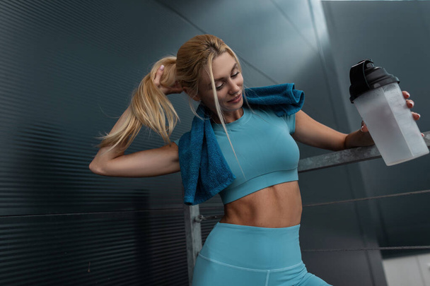 Фитнес-блондинка в модном синем костюме позирует у темной металлической стены в городе - Фото, изображение