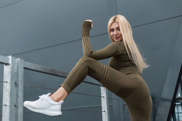 Güzel fitness modeli yeşil spor elbiseli sarışın kız şehirde metal bir duvarın yanında poz veriyor. - Fotoğraf, Görsel