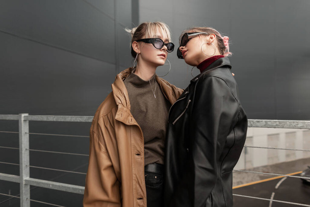 Elegante giovane coppia di belle ragazze con occhiali da sole in giacca di pelle di moda per strada vicino a un edificio moderno - Foto, immagini