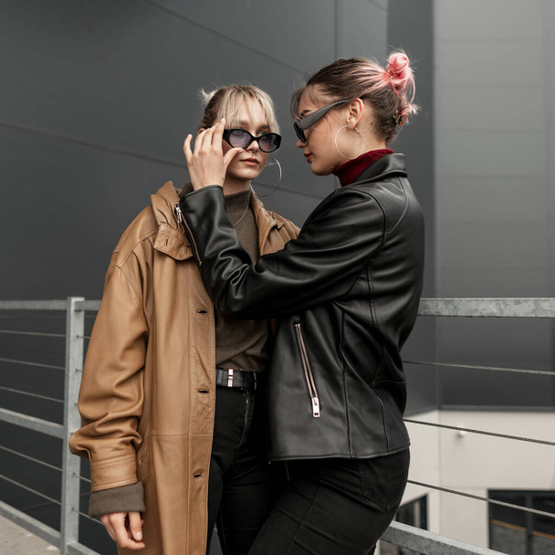 Two beautiful teen sisters girls in a fashionable leather jacket with black jeans in the city. Woman adjusts another woman's sunglasses. - Photo, Image