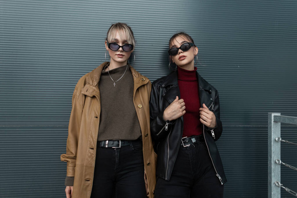 Vogue dwie modne młode kobiety w stylowych okularach przeciwsłonecznych w skórzanych kurtkach z czarnymi dżinsami, pozujące przy metalowej ścianie na ulicy - Zdjęcie, obraz