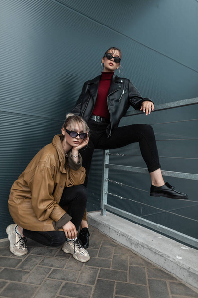 Zwei schöne junge Frauen in trendiger Jeanskleidung mit Lederjacke und Sonnenbrille posieren an einer Metallwand im Freien - Foto, Bild