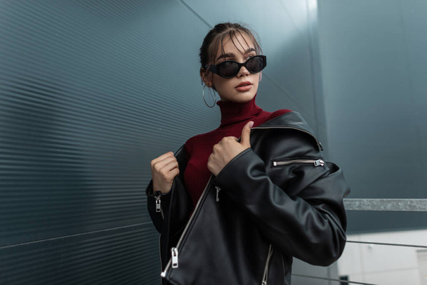 Hermoso futuro modelo de mujer de moda en gafas de sol con estilo con chaqueta de mezclilla negro y suéter de color burdeos posando cerca de una pared de metal en la ciudad moderna - Foto, imagen