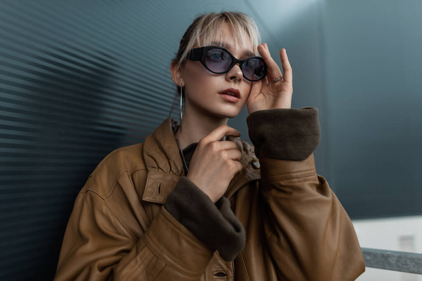 Retrato de moda de uma bela jovem mulher em uma jaqueta de couro marrom vintage com óculos de sol elegantes perto de uma parede de metal preto na cidade - Foto, Imagem