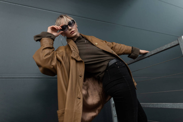 Youth beautiful young woman in fashion sunglasses in stylish brown leather jacket with sweater and black jeans posing near metal wall - Fotoğraf, Görsel