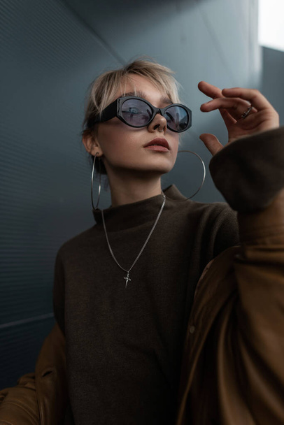 Модный портрет красивой девушки-модели в стильных солнцезащитных очках в коричневой куртке и свитере в городе - Фото, изображение