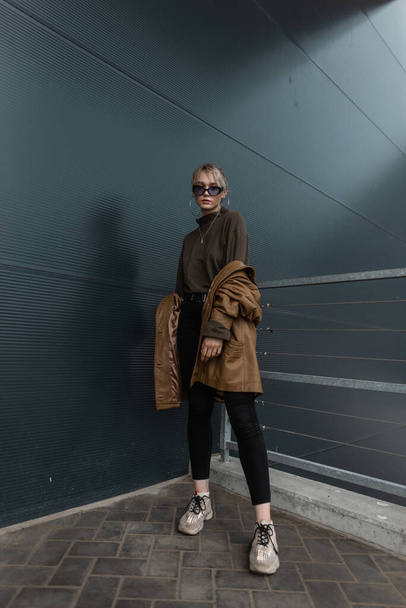 Beautiful young girl model in a fashionable leather jacket with black jeans and sneakers in sunglasses near a metal modern wall in the city - Photo, Image