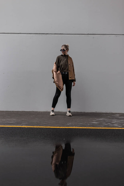 Elegante joven hermosa chica en una chaqueta de cuero marrón de moda con un suéter en vaqueros negros con zapatillas de deporte cerca de una pared gris en la ciudad - Foto, Imagen