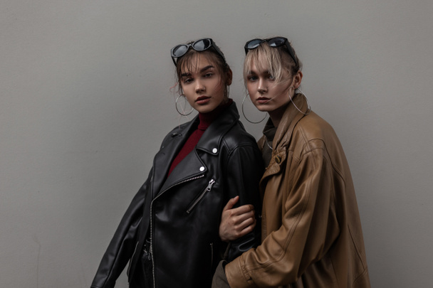 Две красивые молодые модные сестры девушки в модной одежде стоят возле серой стены в городе - Фото, изображение