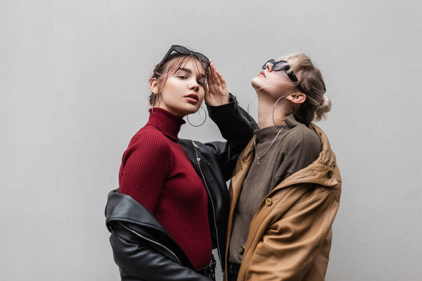 stylish beautiful young girlfriends woman in a fashionable leather jacket and knit sweater with sunglasses stand on a gray background in the city - Foto, imagen