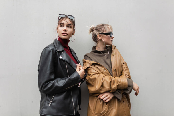 Twee modieuze mooie modelmeisjes in een vintage leren jasje met zonnebril in een trui staan bij een grijze muur in de stad. Jongerenstijl - Foto, afbeelding