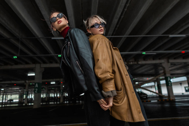 Two fashion models girls in stylish city clothes with a leather jacket with cool sun goggles posing near a parking lot in the city - Photo, Image
