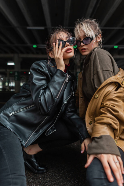 Deux belles filles urbaines élégantes avec des lunettes de soleil de mode et vestes en cuir vintage avec pull posant dans la ville. Style de jeunesse futur - Photo, image