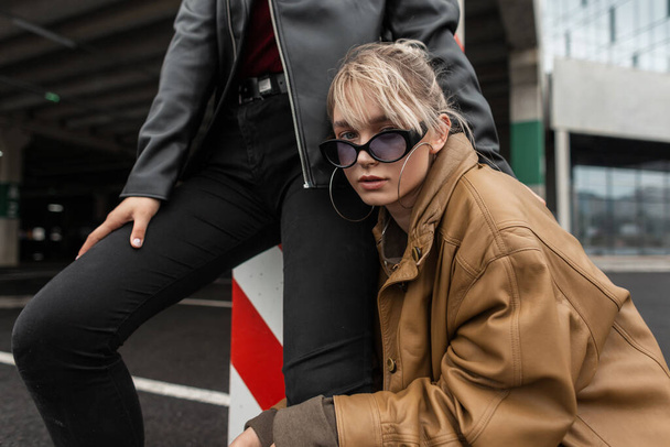 Trendy jonge vogue mooie stijlvolle vrouw met zonnebril en een vintage leren jas poseren in de buurt van een meisje in zwarte jeans in de stad - Foto, afbeelding