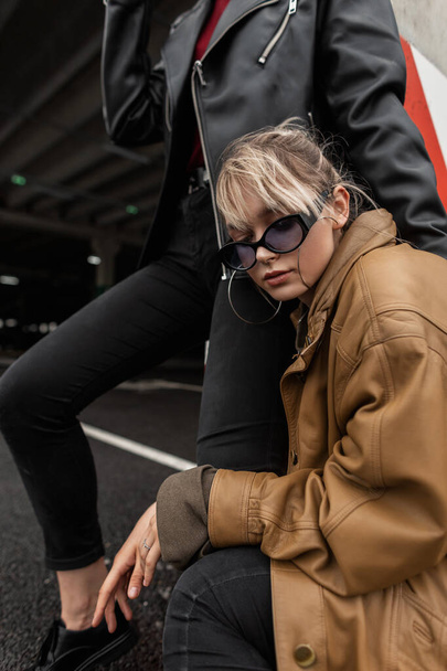 Крутая стильная молодая девушка в старинных солнцезащитных очках в модной коричневой кожаной куртке с модной джинсовой одеждой с подружкой позирует в городе - Фото, изображение