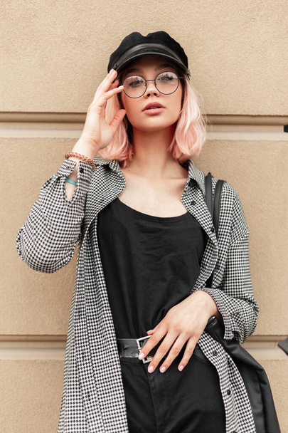 Kaunis malli teini nainen lasit tyylikäs lippis rento ruudullinen paita musta t-paita seisoo lähellä beige seinä kadulla ja katsoo kameraa - Valokuva, kuva