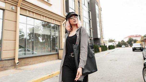 Stilvolle junge schöne Frau mit Brille und Hut in modischer Kleidung mit Hemd und schwarzer Jeans mit Tasche spaziert durch die Stadt - Foto, Bild