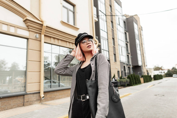 Mujer joven elegante urbana con gafas vintage y una gorra en una camisa a cuadros de moda con una camiseta negra, jeans y una bolsa de moda paseos por la ciudad cerca del edificio - Foto, Imagen