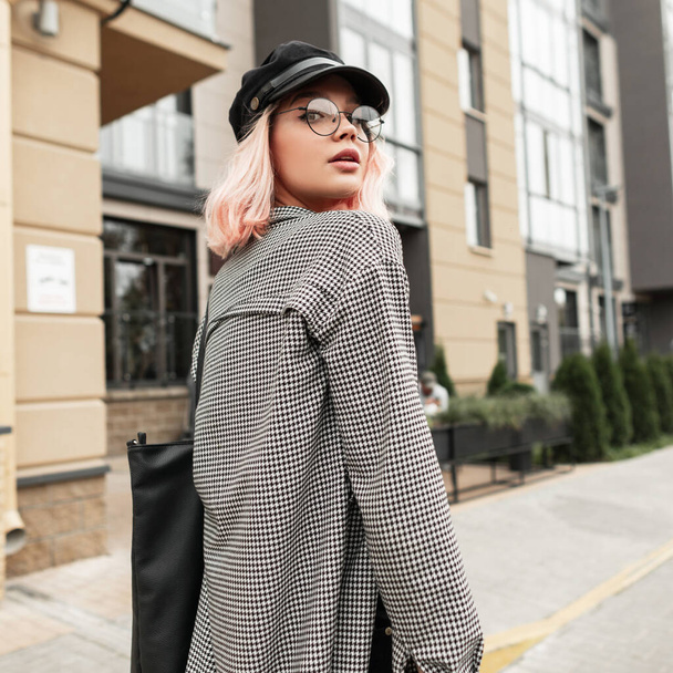 Cute young woman with stylish glasses and vintage hat with trendy casual shirt walking on the street near the building - Фото, изображение