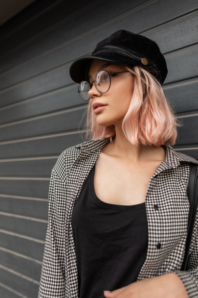 Portrait de la mode jolie femme modèle avec des cheveux roses dans une casquette et chemise décontractée à la mode avec des lunettes se tient près d'un mur gris dans la ville - Photo, image