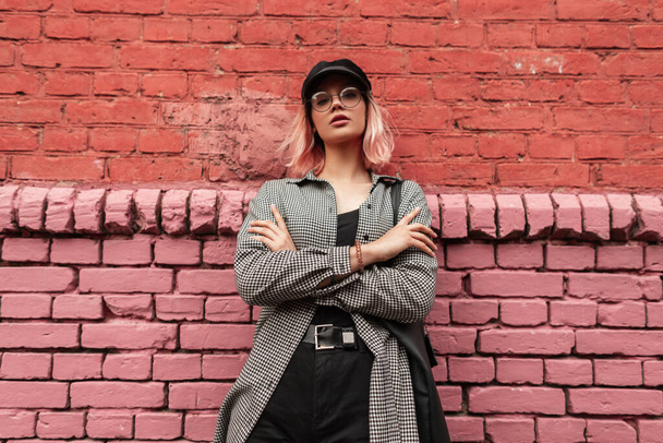 Hipster modelo de mulher jovem elegante com óculos elegantes na camisa xadrez na moda e boné vintage fica perto de tijolo parede vermelha e rosa na rua - Foto, Imagem