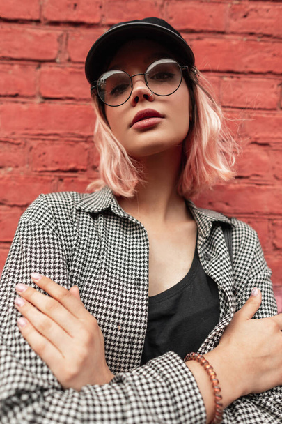 Fashion portrait of a cute young woman with a beautiful face with stylish glasses and a hat in a fashion plaid shirt on the street near a brick wall - Photo, Image