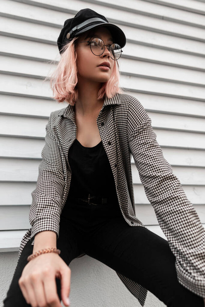 Mulher modelo muito hipster com óculos elegantes e um boné na moda em roupas jeans com uma camisa xadrez fica perto de uma parede de madeira - Foto, Imagem