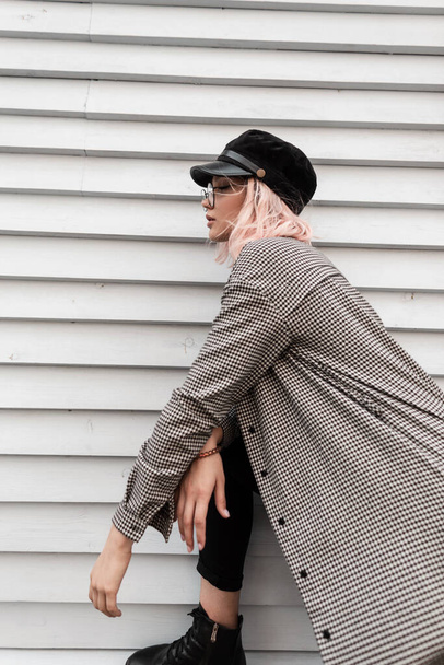 Modelo de mulher bonita elegante com óculos vintage e um boné de moda com uma camisa xadrez casual posando perto de uma parede de madeira na rua - Foto, Imagem