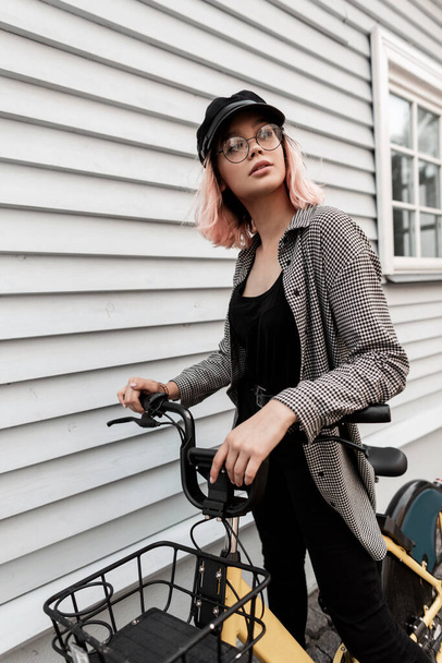 Hipster junge schöne Mädchen in modischen Freizeitkleidung reist und fährt auf einem Fahrrad in der Nähe eines Holzhauses auf der Straße - Foto, Bild