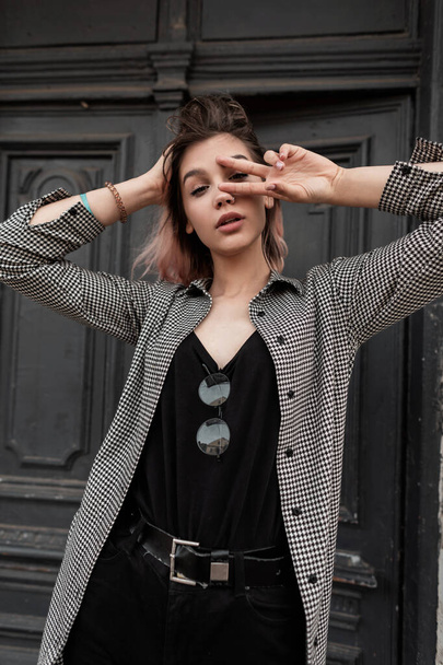 Стильная красивая кавказская хипстерша в модной клетчатой рубашке показывает знак мира на сером винтажном фоне двери на улице - Фото, изображение